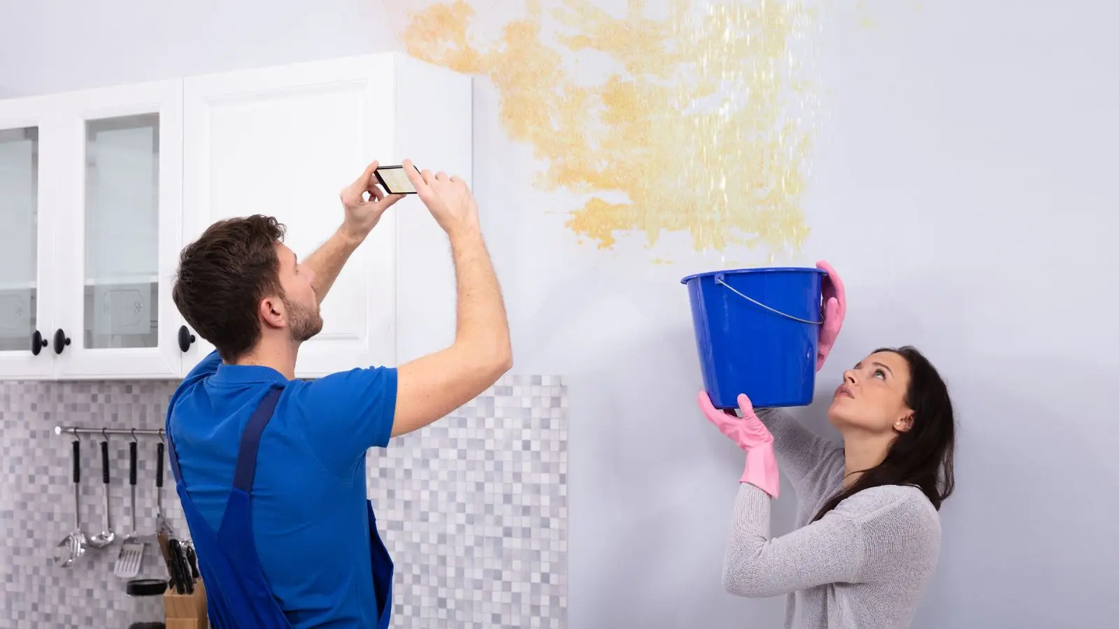 A man photographing a ceiling water stain while a woman holds a bucket under the leak